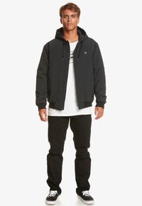 Black zip-up jacket with a hood, paired with a white t-shirt and black pants. Features a small chest logo and side pockets.