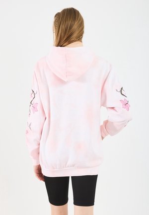 Person iført en lyserød hættetrøje med blomstergrenmotiver på ærmerne, stående med ryggen til, kombineret med sorte cykelshorts.
