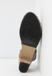 Semelle d’une chaussure à talon avec une semelle en caoutchouc noir texturé et un talon en bois marron clair, positionnée debout contre des murs d’angle blancs.
