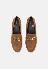 Mocassins en daim marron avec un bout rond, ornés d'un détail en métal doré à l'avant. Semelles intérieures noires avec le logo de la marque visible à l'intérieur.