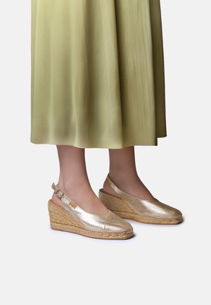 Pies con alpargatas de cuña metálicas doradas con tiras en el tobillo, combinadas con una falda midi plisada de color verde claro.