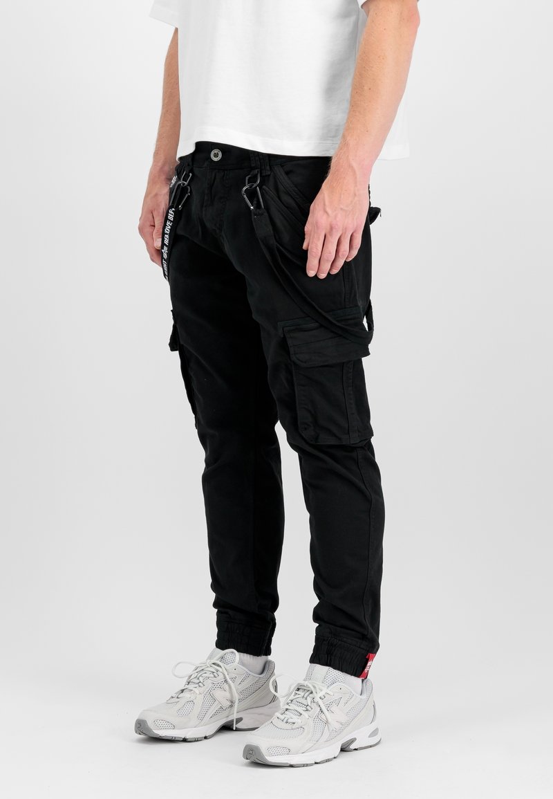 Personne portant un pantalon cargo noir avec poches et détails de sangles, un t-shirt blanc et des baskets de sport blanches, debout devant un fond uni.