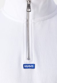 Bílý výrobek s vysokým límcem a stříbrným zipem, pod kterým se nachází malá modrá obdélníková značka s nápisem "HUGO".
