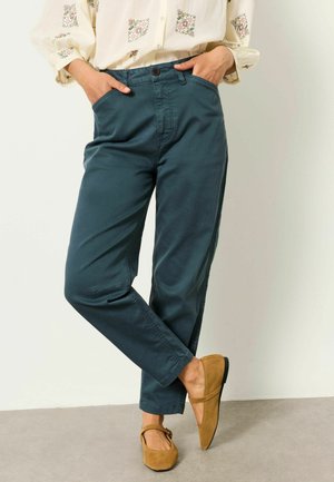 Person trägt dunkelteal-farbene, gerade geschnittene Hose, beige Bluse mit Blumenmuster und senfgelbe Wildleder-Ballerinas, sitzt mit gekreuzten Beinen und Händen in den Taschen.