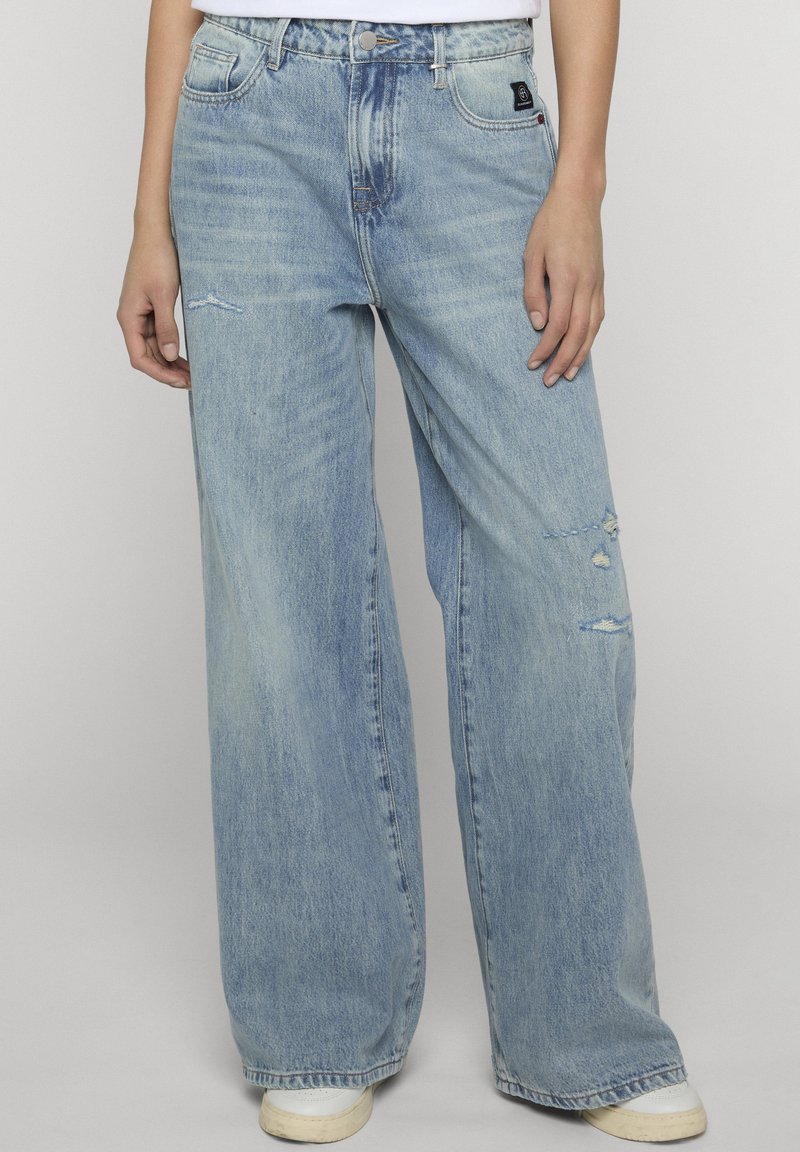 Personne portant un jean large bleu clair avec une légère usure et des baskets blanches, debout devant un fond uni.