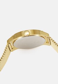 Goldfarbene Uhr mit einem runden Gehäuse, strukturiertem goldenen Mesh-Armband und einer Seitenkrone zur Anpassung. Die Rückseite hat eine glatte silberne Oberfläche.