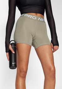 Oliwkowe szorty Nike Pro z szarym pasem. Model trzyma czarną butelkę na wodę Nike, prezentując smukły, sportowy design.