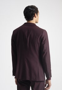 Hombre con cabello corto y rizado de color oscuro visto desde atrás, vestido con una chaqueta de traje burdeos oscuro y pantalones a juego, frente a un fondo blanco.