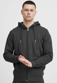 Hoodie zippé gris foncé en tissu doux. Équipé d'une capuche, de cordons de serrage, de poches avant et de poignets côtelés avec des accents en métal argenté.