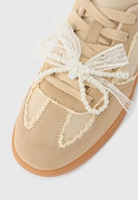 Μπεζ σουέντ αθλητικό παπούτσι με δαντελένιο τελείωμα και διακοσμητικό φιόγκο από λευκές πέρλες και δαντέλα στα κορδόνια.