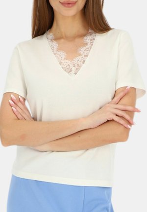 Kobieta w białym topie z koronkowym wycięciem w serek i niebieskich spodniach, z rękami skrzyżowanymi na piersi.