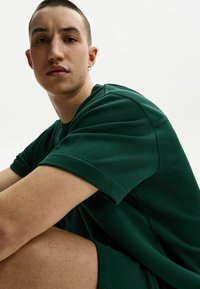 Zöld pamut póló laza fazonnal, rövid ujjakkal és bordázott nyakkivágással, amelyhez illeszkedő sortot visel. Simaság jellemzi, minimalista dizájn.