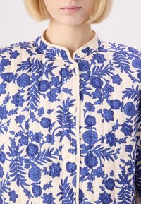 Quilted jacket featuring a cream background with blue floral patterns, round neck, and front buttons. Textured fabric with a smooth finish.