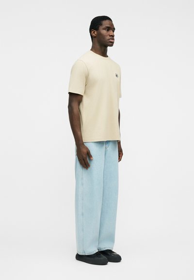 Άνδρας όρθιος, φοράει μπεζ κοντομάνικη μπλούζα, ανοιχτό μπλε τζιν παντελόνι με φαρδιά μπατζάκια και μαύρα αθλητικά, μπροστά σε απλό λευκό φόντο.