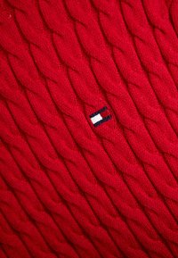 Tissu en maille torsadée épais rouge avec un petit logo rectangulaire présentant des rayures horizontales blanches, rouges et bleu marine.