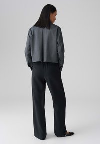 Grijze kort gerdenk jacket met een gladde textuur en subtiele verticale strepen, gecombineerd met losse zwarte broek en zwarte flats.