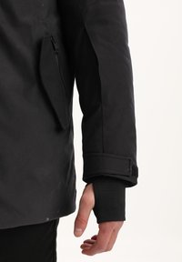 Giacca nera da esterno con tasca zip sulla manica, finitura testurizzata e regolazione con velcro sui polsini. Mano visibile che indossa una manica nera.