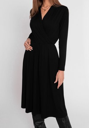 Vrouw die een zwarte midi-jurk met lange mouwen draagt, met een V-hals en overslag aan de voorkant, gecombineerd met zwarte kniehoge laarzen.