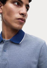 Polo bleu avec un col bleu foncé et des accents blancs. Tissu texturé, patte de boutonnage à deux boutons et manches courtes.
