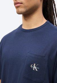 Άνδρας με μακριά μαλλιά φοράει μπλε ναυτικό T-shirt Calvin Klein με τσέπη στο στήθος και λογότυπο, εμφανίζεται από το στήθος και πάνω σε λευκό φόντο.