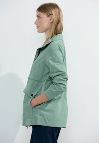 Chaqueta cortavientos de color verde claro con cremallera en la parte delantera, cuello doblado y gran bolsillo frontal. Cuenta con un parche de la marca en la manga izquierda. Ajuste regular.