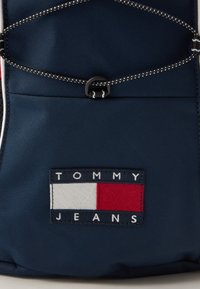 Modrá nylonová batoh s výrazným vyšitým logom "TOMMY JEANS" s červenými, bielymi a modrými farebnými blokmi a čiernymi šnúrkami.