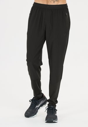 Homme portant un jogger de sport noir avec des poches zippées et des chaussures de course noires, debout sur un fond clair uni.