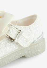 Scarpa bianca glitterata con un grande fiocco crema, fibbia argentata e suola trasparente texture. La superficie scintilla con piccole paillettes.