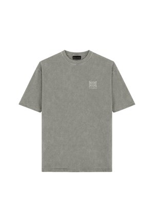 Man staat met het gezicht naar voren, draagt een verbleekte groene oversized T-shirt, donkergrijze spijkerbroek en zwart-witte sneakers tegen een eenvoudige achtergrond.