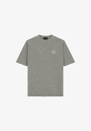 Mann steht frontal, trägt ein verblasstes grünes, übergroßes T-Shirt, dunkelgraue Jeans und schwarz-weiße Sneakers vor schlichtem Hintergrund.
