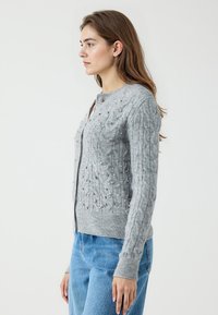 Grijze gebreide cardigan met een textuur kabelpatroon en voor versieringen. Heeft een ronde halslijn en ribbelmanchetten en -zoom.