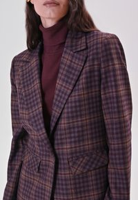 Eine Frau trägt ein lila karierter Blazer über einem passenden lila Rollkragenpullover, mit sichtbaren langen dunklen Haaren und einem neutralen Lippenfarbton.