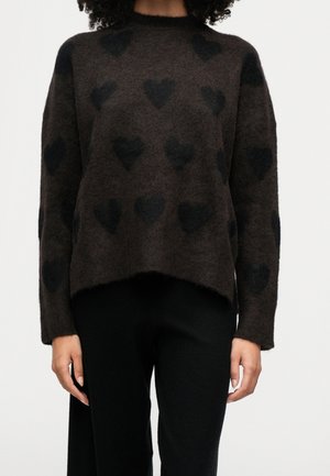 Modèle portant un pull marron foncé à motifs de cœurs noirs et un pantalon noir, sur un fond uni clair.