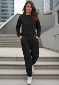 Fekete pulóver, kerek nyakkivágással és raglán ujjal, fekete sportos nadrággal párosítva, amely rugalmas derékrésszel és oldalsó zsebekkel rendelkezik. Fehér sportcipő.