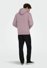Man die een mauve hoodie draagt met de capuchon naar beneden, zwarte spijkerbroek en zwarte sneakers, staand met handen in de zakken van de hoodie, met de rug naar de camera.