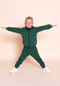 Zielony bawełniany dres z rozpinaną kurtką na zamek i spodniami z elastycznymi ściągaczami. Dodatki to naszywka z logo i kontrastowe lamówki. Białe trampki.