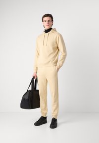 Hanorac bej și pantalonii asortați dintr-un material moale. Adidași negri și o geantă de sport neagră. Design simplu, croială relaxată și accente funcționale.