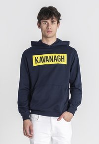 Navy modrá mikina s kapucňou vyrobená z bavlneného zmesi, s veľkým žltým logom "KAVANAGH" vpredu a klokanie vrecko. Rúbkované manžety a lem.
