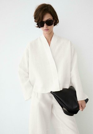 Femme aux cheveux bruns courts portant de grandes lunettes de soleil noires, un haut blanc ample et un pantalon, tenant une grande pochette en cuir noir.