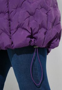 Lila gesteppte Jacke mit Kordelzugsaum über blauen Jeans an einer Person vor einem hellen Hintergrund.