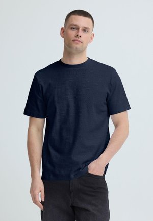 Mlad moški s kratkimi lasmi, oblečen v teksturirano temno modro srajco s kratkimi rokavi in črne hlače, stoji z eno roko v žepu.