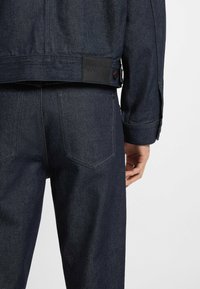Giubbotto in denim scuro e pantaloni, con una finitura testurizzata. Presenta un'etichetta nera sul retro e dettagli con bottoni sulle maniche e sui pantaloni.