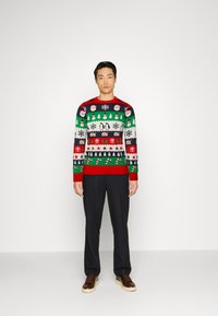 Színes karácsonyi pulóver ünnepi motívumokkal, beleértve a Mikulást, pingvineket és ajándékokat, piros gallérral és szegéllyel. Sötét nadrággal viselve.