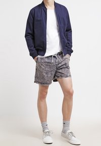 Veste de bombardier marine, t-shirt blanc, shorts gris à motifs, baskets blanches et chaussettes grises. Tenue décontractée avec des textures et des couleurs variées.