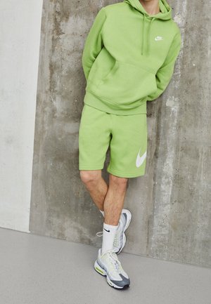 Persoon die een lichtgroene Nike-hoodie en bijpassende korte broek draagt, witte Nike-sokken en witte en grijze sneakers, staand tegen een betonnen muur.
