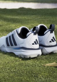 Chaussures de sport blanches et noires avec des crampons reposant sur de l'herbe verte à côté de quelques tees de golf en bois.