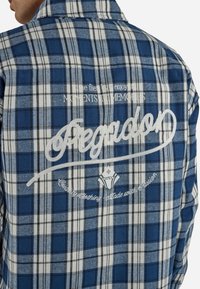 Personne portant une chemise à carreaux bleue et blanche avec un texte brodé blanc indiquant "Pegador" et de plus petites phrases décoratives dans le dos.