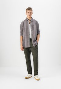 Jovem de pé, vestido com uma camisa quadriculada sobre uma t-shirt branca, calças verde-escuro e ténis brancos, em frente a um fundo branco liso.
