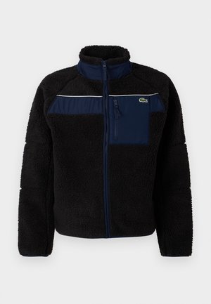 Black fleece jacket with navy accents, high collar, zippered chest pocket, and white piping across the chest. Textured, warm fabric.