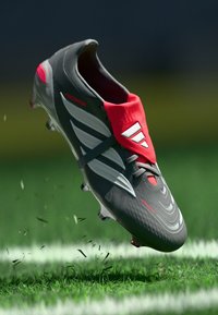 Crampons de football gris avec des accents rouges et des rayures blanches, présentant une tige texturée et un design léger, flottant au-dessus de l'herbe.